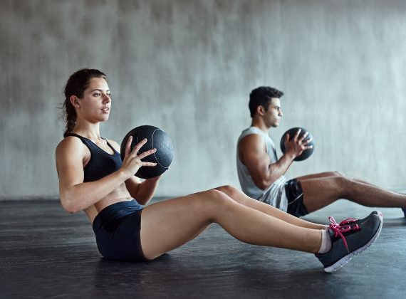Two people engaging in a core workout, utilizing a heavy black gym ball for conditioning exercises.